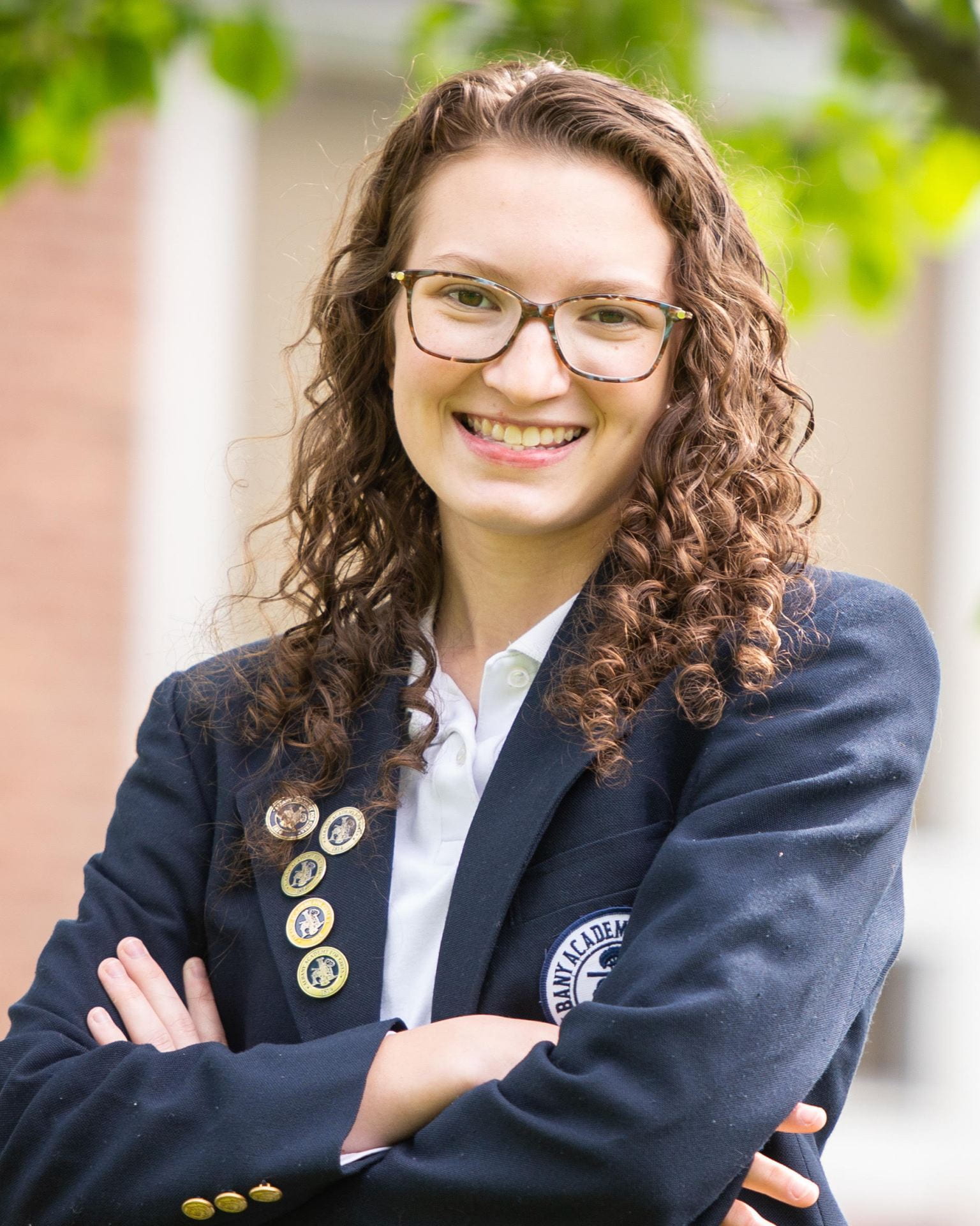 A student with glasses, long, brown curly hair, and a navy blazer, smiles at the camera with her arms crossed.